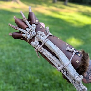 May include: A brown leather gauntlet featuring white bone-like accents shaped like fingers and a forearm plate. The piece is secured with brass rivets and light-colored cord lacing, with dark brown faux fur trim visible at the wrist opening.
