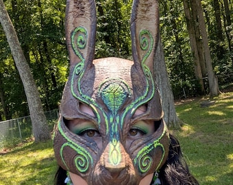 May include: A brown rabbit mask featuring tall, pointed ears and intricate, glowing green swirl patterns across the forehead, cheeks, and ears. The mask has large eye openings and a textured, sculpted surface that covers the upper face.