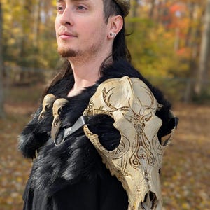 Könnte beinhalten: Ein Kostüm mit einer schulterlangen Knochenschädel-Schulterpartie mit geätzten Designs, einem schwarzen Pelzkragen und einem passenden Kronenaufsatz. Das Schulterstück aus Schädelknochen ist mit einem schwarzen Lederriemen befestigt. Der Hintergrund ist ein verschwommener Herbstwald.