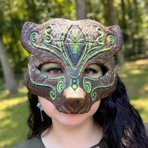 Puede incluir: Una máscara con forma de animal de color marrón con superficies texturizadas y patrones decorativos de volutas en verde brillante. La máscara tiene orejas puntiagudas, un diseño central en la frente y amplias aberturas oculares.