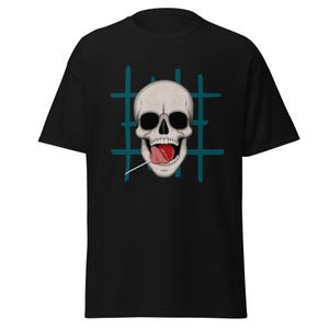 Puede incluir: Camiseta negra con un gráfico de calavera y una piruleta roja. La calavera tiene cuencas de ojos negras y una boca muy abierta con una lengua roja. Un patrón de cuadrícula turquesa está detrás de la calavera.