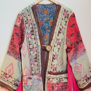 May include: A long, open-front coat with a patchwork design. The coat features a mix of floral and geometric patterns in shades of red, pink, brown, and green. It has a large wooden button closure and bright pink fringe accents.