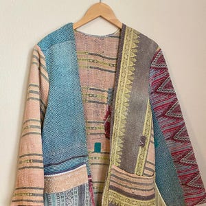 Può includere: Una giacca lunga aperta sul davanti con un design patchwork realizzato in vari tessuti nei toni del rosa, blu, rosso e grigio. Il capo presenta maniche lunghe, due tasche frontali e una decorazione con frange sul bordo inferiore delle maniche.