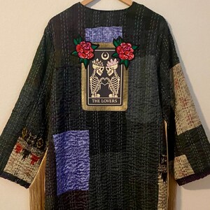 May include: Dark green patchwork kimono with a tarot card patch featuring skeletons and the text "THE LOVERS." The kimono has gold fringe and is hanging on a wooden hanger. Patches of blue and tan fabric are sewn into the design.
