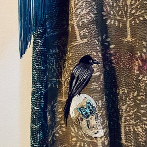 May include: A textured olive green garment featuring a repeating tree pattern, accented by a long blue fringe on the side. An embroidered patch depicts a black crow perched atop a human skull, which is adorned with a small, bright blue butterfly.