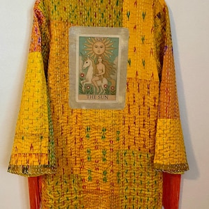 Op de afbeelding: Een lange, gele patchworkjas met een tarotkaart met een zon, een vrouw en een eenhoorn. De jas heeft lange mouwen, oranje franjes en een verscheidenheid aan kleurrijke gestikte patronen.