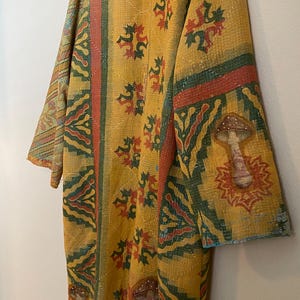 May include: A long-sleeved yellow duster coat featuring a repeating pattern of green and red geometric shapes and floral motifs. The garment is embellished with three-dimensional fabric mushroom appliqués on the sleeves and lower front panels.