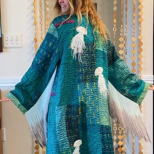 May include: A long, teal-colored patchwork coat featuring a quilted texture and various shades of blue and green. The garment includes three white, jellyfish-shaped fringe appliqués on the back and long, flowing fringe along the sleeves.