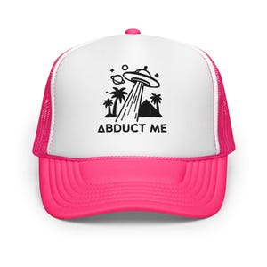 Peut inclure: Casquette de camionneur avec un panneau avant blanc et une visière et un dos en filet rose vif. Le chapeau présente un graphisme noir d'un OVNI projetant sur des palmiers et une pyramide, avec le texte "ABDUCT ME" en dessous.