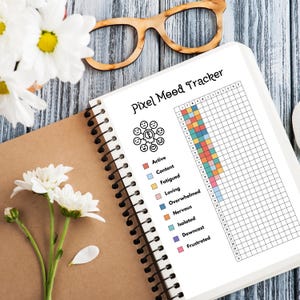 May include: A white notebook with a black spiral binding. The notebook has a black and white pixel mood tracker on the page. The tracker has a grid with different colored squares representing different moods, such as active, content, fatigued, loving, overwhelmed, nervous, isolated, downcast, and frustrated. The notebook is on a wooden surface with a pair of brown glasses and white flowers.