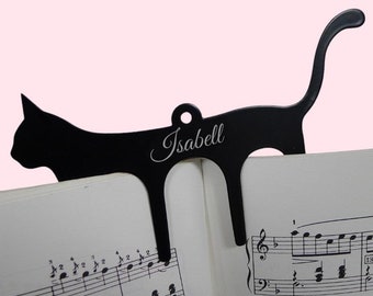 May include: A black metal cat-shaped bookmark with the name "Isabell" in cursive script. The bookmark is placed on a book with musical notes, with the cat's tail curving upwards. The background is a soft pink.