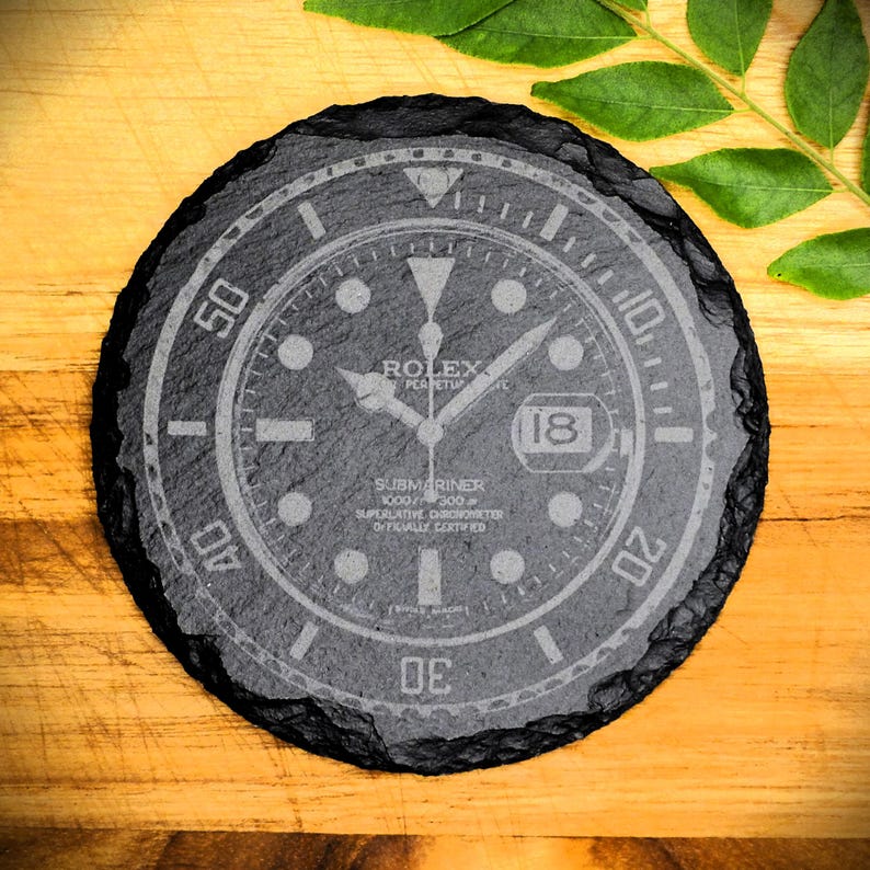 K&ouml;nnte beinhalten: Ein schwarzer Schieferuntersetzer mit einem wei&szlig;en, gravierten Design eines Rolex Submariner Zifferblatts. Das Zifferblatt zeigt den Text "Rolex Perpetual", "Submariner 1000ft=300m", "Superlative Chronometer Officially Certified" und "18".