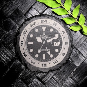 K&ouml;nnte beinhalten: Ein schwarzer und wei&szlig;er Schieferuntersetzer mit einem Rolex Explorer II-Zifferblattmuster. Das Zifferblatt der Uhr zeigt den Text "ROLEX" und "OYSTER PERPETUAL DATE EXPLORER II".