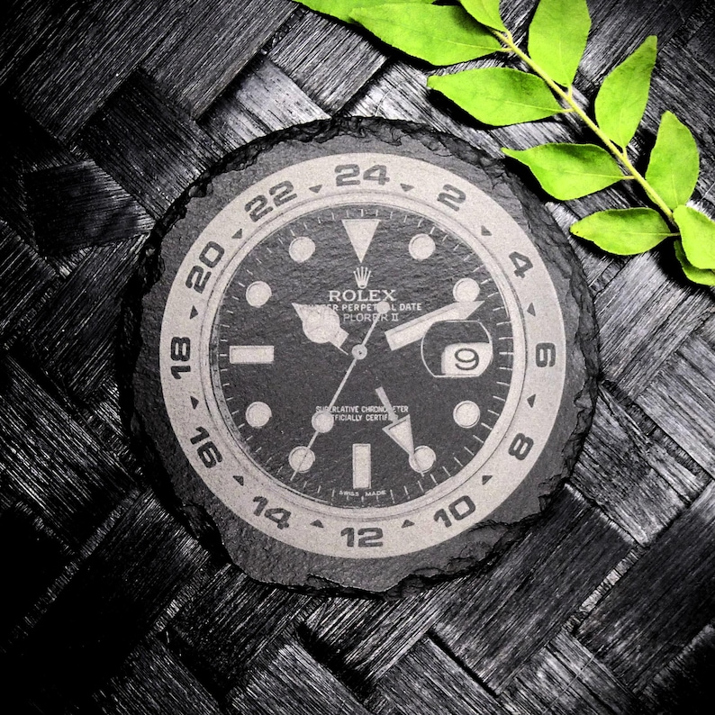 K&ouml;nnte beinhalten: Ein schwarzer und wei&szlig;er Schieferuntersetzer mit einem Rolex Explorer II-Zifferblattmuster. Das Zifferblatt der Uhr zeigt den Text "ROLEX" und "OYSTER PERPETUAL DATE EXPLORER II".