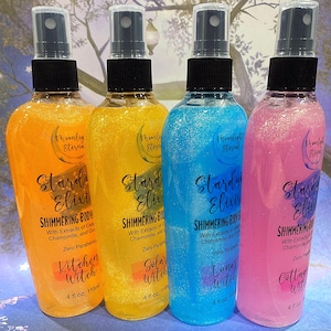 May include: Four shimmering body elixirs in orange, yellow, blue, and pink bottles. Each bottle has a black spray top and contains a glittery liquid. The labels read "Stardust Elixir" and feature the names "Kitchen Witch," "Solar Witch," "Lunar Witch," and "Cottage Witch."