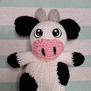 May include: A white knitted cow plush toy with black spots and a pink nose. The cow has large black eyes with white stars.