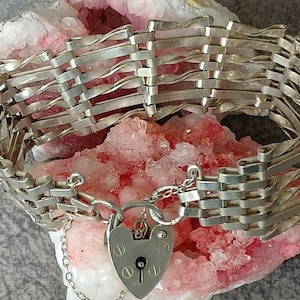 Könnte beinhalten: Ein silberfarbenes Armband mit gewebtem Design. Das Armband hat einen herzförmigen Vorhängeschlossverschluss und eine zarte Kette. Der Schmuck wird auf einer rosa-weißen Kristallformation präsentiert.