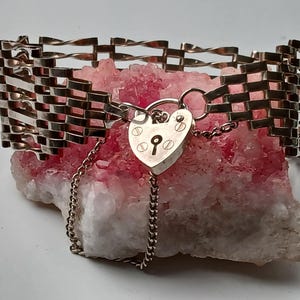 Puede incluir: Una pulsera plateada con un diseño tejido, con un cierre de candado en forma de corazón y una delicada cadena. La pulsera se exhibe sobre una formación de cristal rosa y blanco, mostrando sus intrincados detalles y estética vintage.