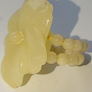 May include: A light yellow ring with a carved floral design and a beaded band. The floral element has a smooth, glossy finish, and the band is made of small, round beads. This unique ring is a piece of jewellery.