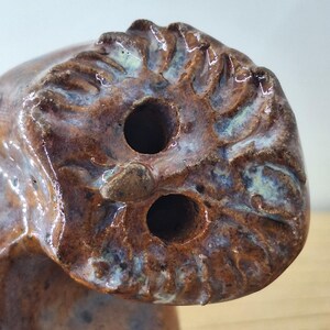May include: A brown ceramic object with a glossy finish and two circular holes on its textured, uneven surface. The glaze features subtle blue and white highlights within the crevices of the moulded, organic-looking clay structure.