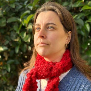 May include: A woman wearing a bright red, textured scarf and a blue sweater. The scarf is wrapped around the neck and contrasts with the blue of the sweater. The background is a blurred view of green foliage.
