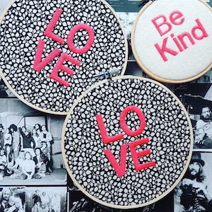 Könnte beinhalten: Gestickte Reifen mit dem Wort "LOVE" in leuchtend rosa Garn auf einem schwarz-weiß gemusterten Stoff. Ein kleinerer Reifen zeigt die Worte "Be Kind" in rosa Garn auf weißem Hintergrund. Die Reifen werden vor einem Hintergrund aus Schwarz-Weiß-Fotografien ausgestellt.