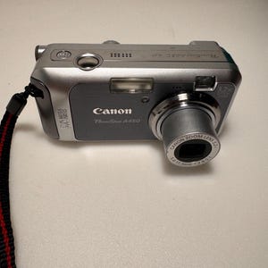 May include: A silver and grey Canon PowerShot A450 digital camera with a 3.2x optical zoom lens. The front features the Canon logo, 5.0 megapixels text, and a built-in flash. A black wrist strap with a red stripe is attached to the side of the device.