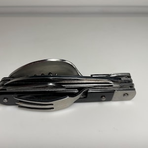 Puede incluir: Una multiherramienta con tenedor, cuchara, sacacorchos y otros utensilios. La herramienta está hecha de acero inoxidable y plástico negro. El tenedor mide aproximadamente 3,8 cm de ancho.
