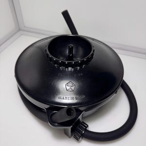 May include: A black circular device with a central knob and a flexible black hose attached to the side. The top surface features the text "MADE IN USSR" and a logo with the letters "CCCP" inside a pentagon shape, along with other markings.