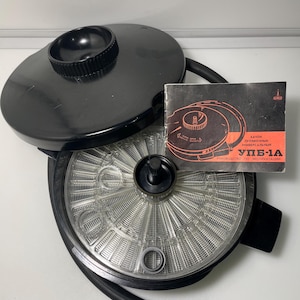 Może przedstawiać: Czarny zbiornik do wywoływania filmów z przezroczystym, okrągłym wnętrzem i czarną pokrywą. Mała książeczka z instrukcjami z czerwonym i czarnym tekstem znajduje się na górze zbiornika. Zbiornik ma dołączoną czarną gumową rurkę.