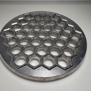以下が含まれることがあります： 銀色の金属製で円形の餃子成形器。生地を成形するための複数の開いた円形の穴が、六角形のグリッドパターンで繰り返し配置されています。このツールには平らな外縁があり、表面全体に質感のある仕上げが施されています。