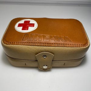 Può includere: Un kit di pronto soccorso in pelle color cuoio con una croce rossa e la parola "АПТЕЧКА" in rilievo sul davanti.