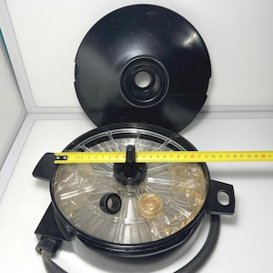 Puede incluir: Una vaporera eléctrica negra con un interior transparente y circular y una tapa negra. El diámetro de la vaporera es de aproximadamente 25 cm. Una cinta métrica amarilla se coloca sobre la vaporera.