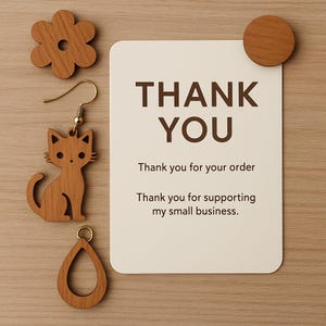May include: Wooden cat-shaped earring, teardrop-shaped earring, and flower-shaped wooden accent pieces. A white card with the words "THANK YOU" and two lines of text is attached to a wooden surface.