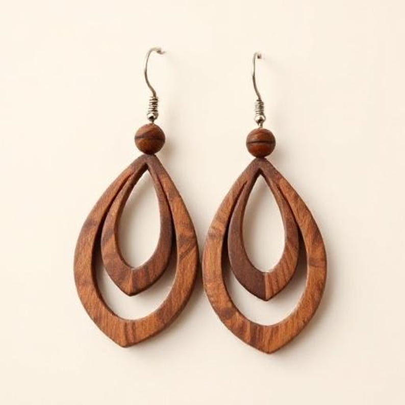 May include: A pair of wooden teardrop earrings. Each earring features a layered design with a small wooden bead at the top and a silver-colored hook. The earrings are a warm brown color and have a natural wood grain.