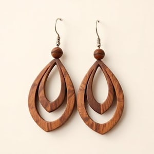 May include: A pair of wooden teardrop earrings. Each earring features a layered design with a small wooden bead at the top and a silver-colored hook. The earrings are a warm brown color.