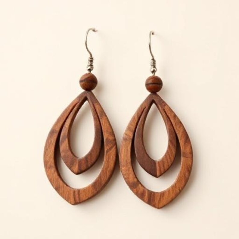 May include: A pair of wooden teardrop earrings. Each earring features a layered design with a small wooden bead at the top and a silver-colored hook. The earrings are a warm brown color.