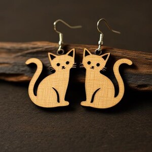 May include: A pair of wooden cat-shaped earrings. The earrings are light brown with black details for the eyes, nose, and mouth. The cat's tail is curled. The earrings hang from silver-colored hooks.