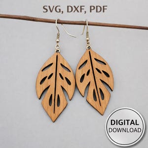 Puede incluir: Un par de pendientes de madera con forma de hoja con detalles recortados, colgando de ganchos plateados. Los pendientes son de color marrón claro y se muestran sobre un fondo neutro. La imagen también incluye el texto "SVG, DXF, PDF" y una insignia de "DESCARGA DIGITAL".