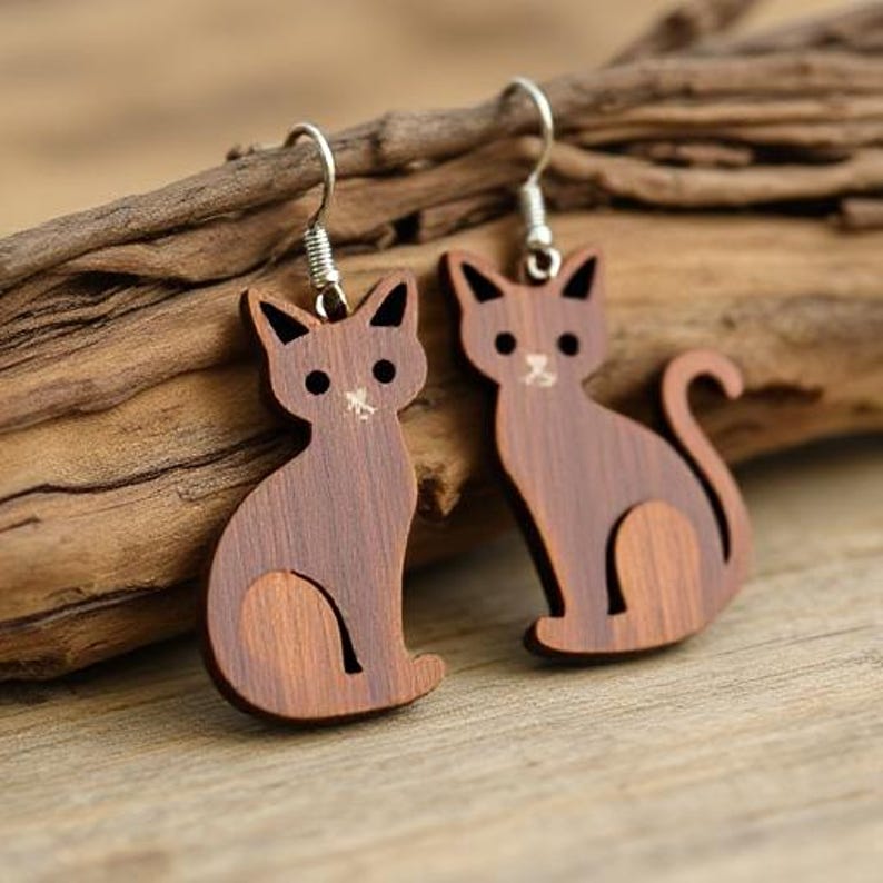 May include: A pair of wooden cat earrings. Each earring is a silhouette of a sitting cat with a curved tail. The earrings are brown with black accents and silver-colored hooks. The earrings are hanging on a piece of wood.