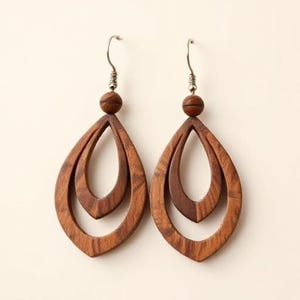 May include: A pair of wooden teardrop earrings. Each earring features a layered design with a small wooden bead at the top and a silver-colored hook. The earrings are a warm brown color.