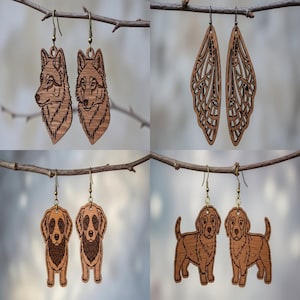 May include: Four pairs of wooden earrings are displayed. Two wolf head earrings, two butterfly wing earrings, two dog head earrings, and two dog body earrings. All earrings are brown with brass-colored hooks, hanging from a tree branch.