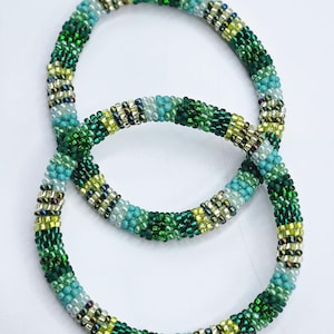 May include: Two beaded bracelets in shades of green, teal, and yellow. The bracelets are made of small, colorful beads and are arranged in a figure-eight pattern against a white background. The bracelets are likely handmade.