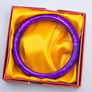 May include: A vibrant purple bangle bracelet with a marbled pattern, presented in a red box lined with golden yellow satin. The bracelet is a circular shape and appears to be made of stone or a similar material.