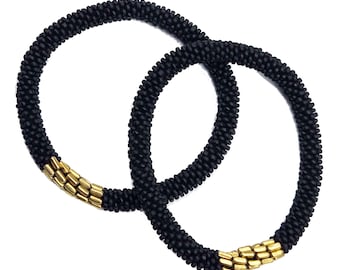 May include: Two black beaded bracelets with gold-coloured accents. The bracelets are circular and appear to be made of small, tightly packed beads. The gold accents are rectangular and placed at intervals around the bracelets.