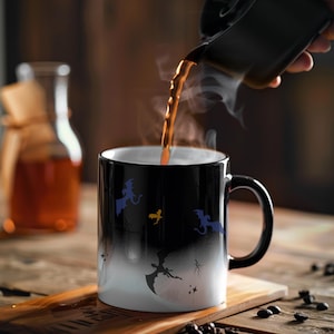 Op de afbeelding: Een zwarte hittegevoelige mok met een ontwerp van draken en een maan die verschijnt wanneer er hete vloeistof in de mok wordt gegoten. De mok staat op een houten oppervlak met koffiebonen eromheen.
