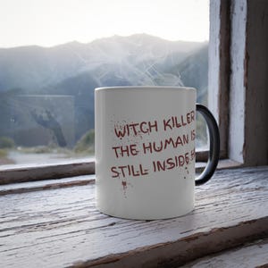Könnte beinhalten: Eine weiße Keramiktasse mit schwarzem Henkel, mit dem Text "WITCH KILLER THE HUMAN IS STILL INSIDE." in blutroter Schrift. Dampf steigt aus der Tasse auf, was auf ein heißes Getränk hindeutet.