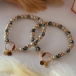 Könnte beinhalten: Zwei Hunde-Halsbänder mit Perlen. Jedes Halsband hat cremefarbene Perlen, goldfarbene Perlen und schwarz-weiß gemusterte Perlen. Goldfarbene Metallringe und Pfotenabdruck-Anhänger vervollständigen das Design. Die Halsbänder werden auf einer weißen Oberfläche präsentiert.