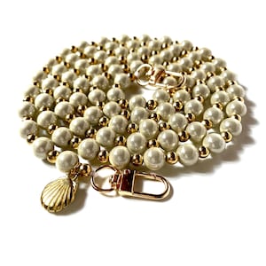 Peut inclure: Une chaîne en perles blanches et dorées avec un charme en forme de coquillage doré et deux fermoirs dorés.