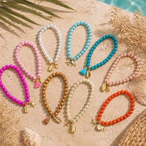 Puede incluir: Una colección de nueve pulseras de cuentas en varios colores, incluyendo rosa, blanco, azul claro, turquesa, bronce, crema y naranja. Cada pulsera presenta un pequeño dije dorado con forma de concha o estrella de mar, y algunas incluyen borlas.